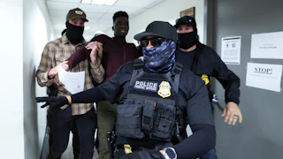 Two masked federal agents detain a man in the hallways of the Jacob K. Javitz Federal Building while another masked agent wearing a flak jacket and gloves walks ahead of them.