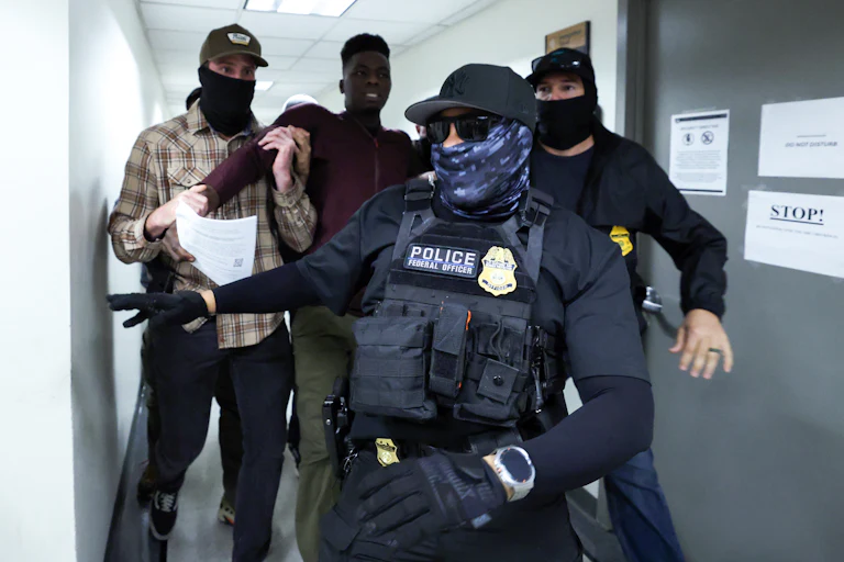 Two masked federal agents detain a man in the hallways of the Jacob K. Javitz Federal Building while another masked agent wearing a flak jacket and gloves walks ahead of them.