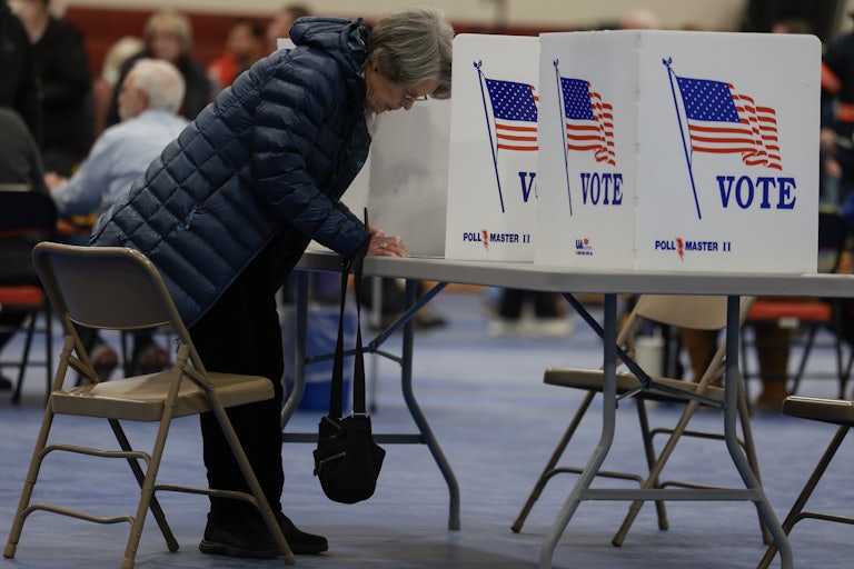 An older woman is bending over and writing something at a table with the "Vote" dividers used at polls.