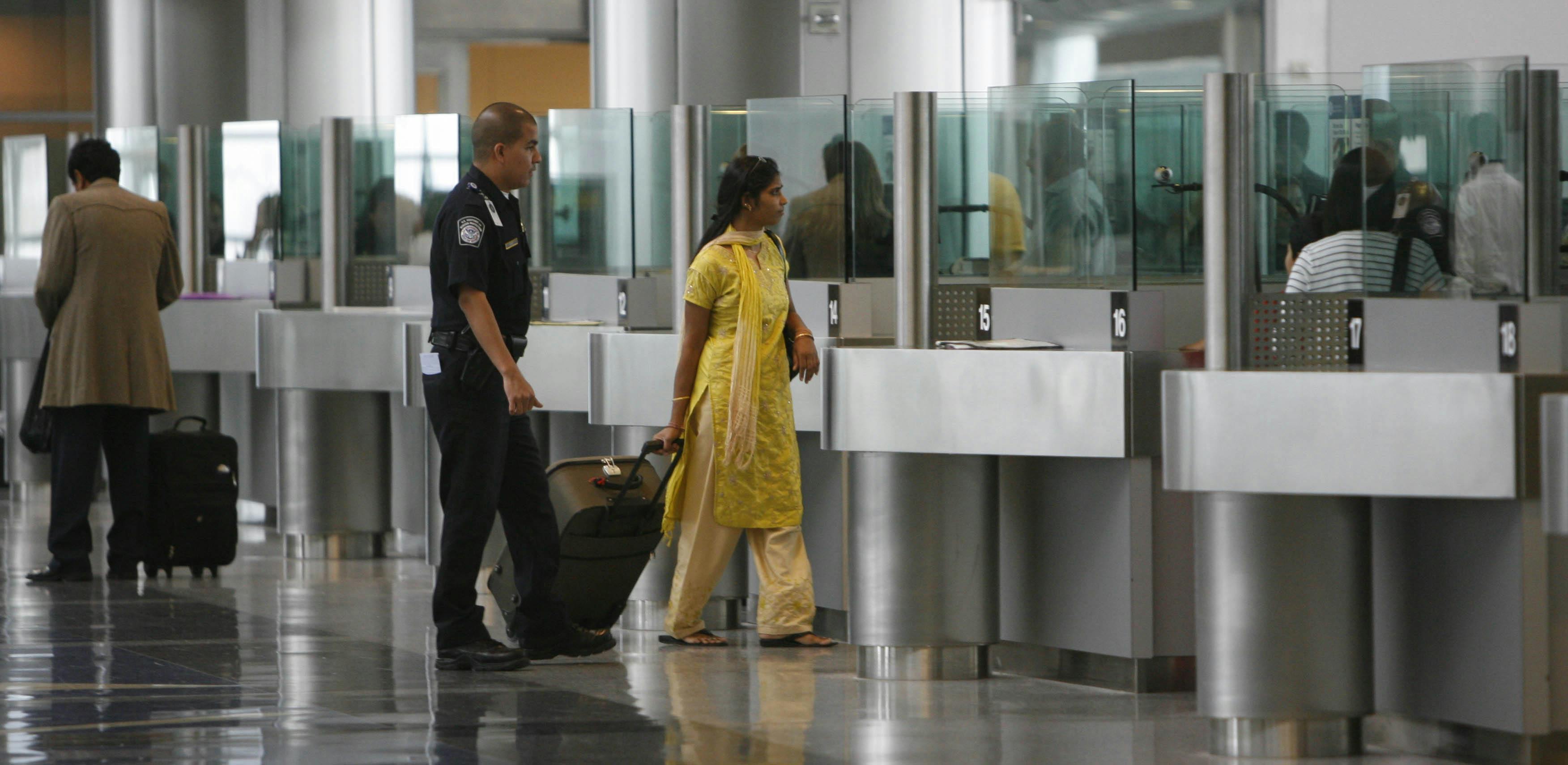 A South Asian woman wearing shalwar kameez walks in an airport with her luggage alongside a police officer.