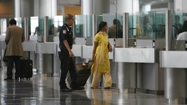 A South Asian woman wearing shalwar kameez walks in an airport with her luggage alongside a police officer.