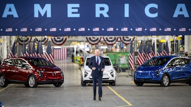 President Biden walks in front of red, white, and blue cars.