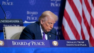 Donald Trump in a seated position, with his head drooping with a blurry American flag behind him to his left and a Board of Peace backdrop behind him.