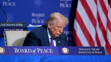 Donald Trump in a seated position, with his head drooping with a blurry American flag behind him to his left and a Board of Peace backdrop behind him.