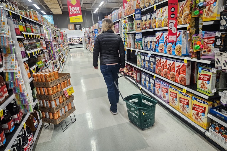 A person pulls a wheeled basket behind them in a grocery store aisle