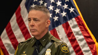 Gregory Bovino stands in uniform angled to the left with an American flag visible beside him.