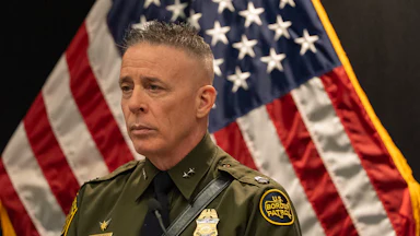 Gregory Bovino stands in uniform angled to the left with an American flag visible beside him.