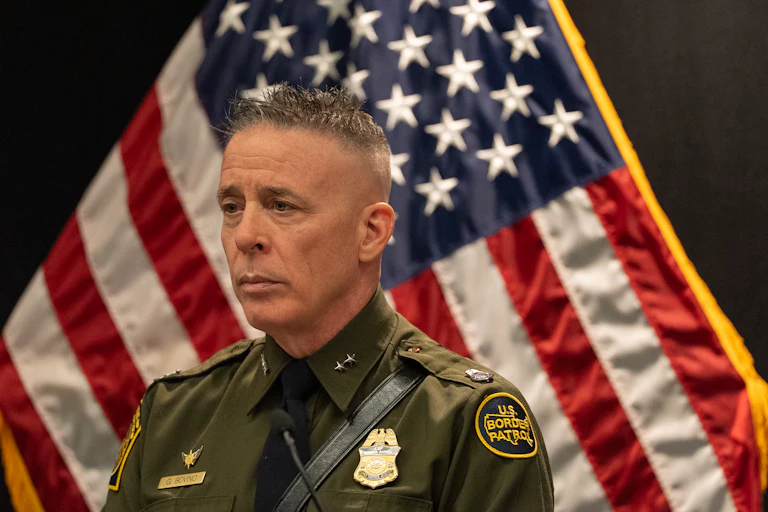 Gregory Bovino stands in uniform angled to the left with an American flag visible beside him.