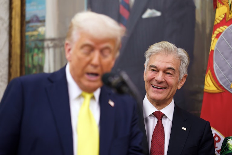 Dr. Mehmet Oz smiles while standing behind Donald Trump, who speaks at a podium in the Oval Office