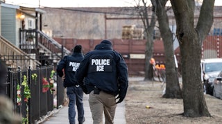 Two ICE agents walk down a neighborhood sidewalk with Christmas deocrations.