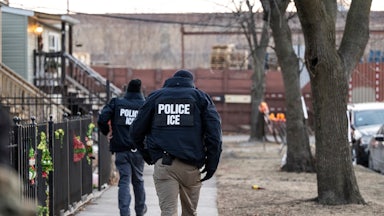 Two ICE agents walk down a neighborhood sidewalk with Christmas deocrations.