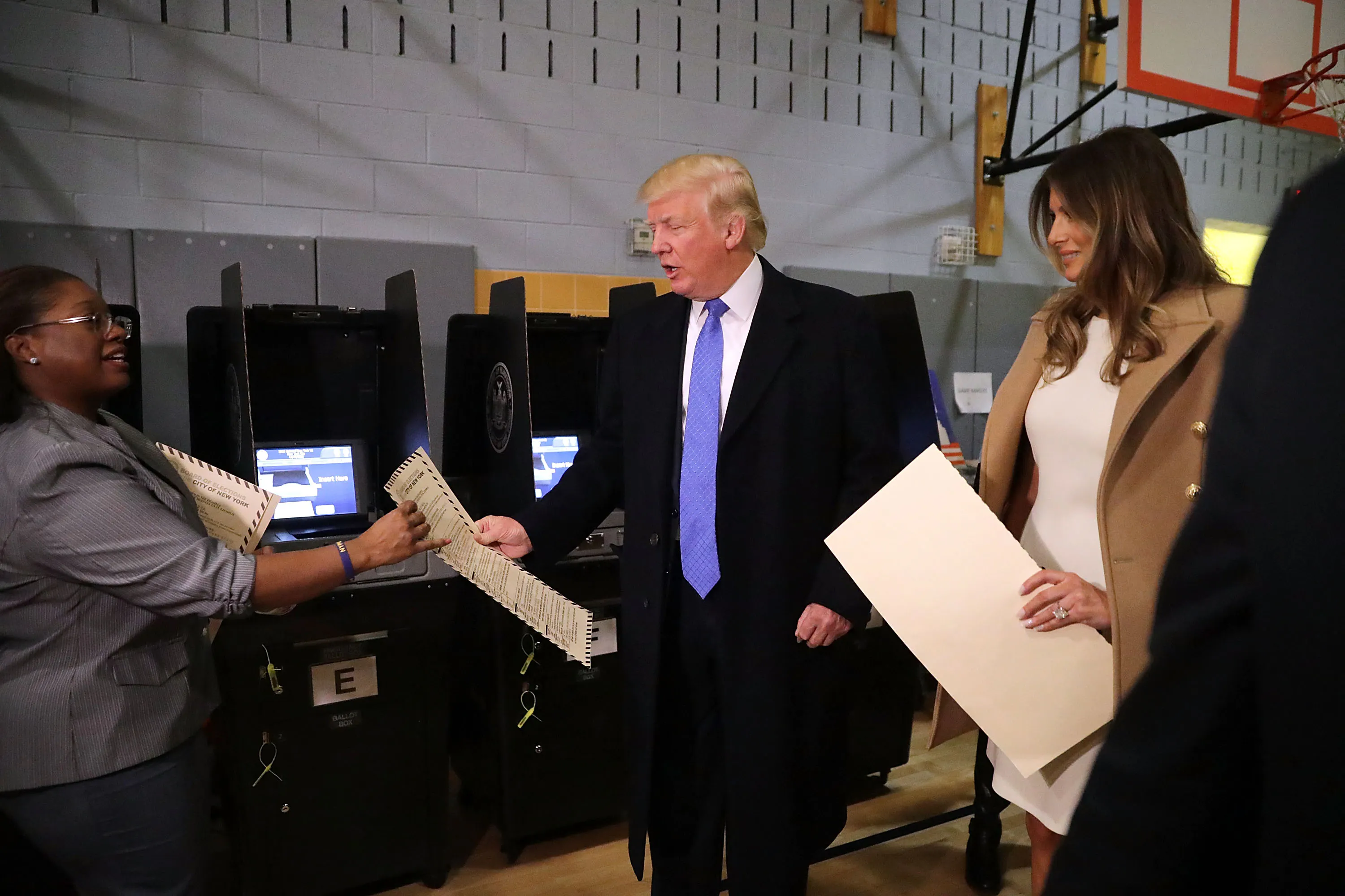 Donald Trump, center, and his wife Melania, right, hand ballots to a poll worker, left in a recreational facility with a basketball hoop visible in the background. 