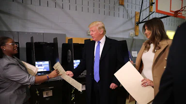 Donald Trump, center, and his wife Melania, right, hand ballots to a poll worker, left in a recreational facility with a basketball hoop visible in the background.