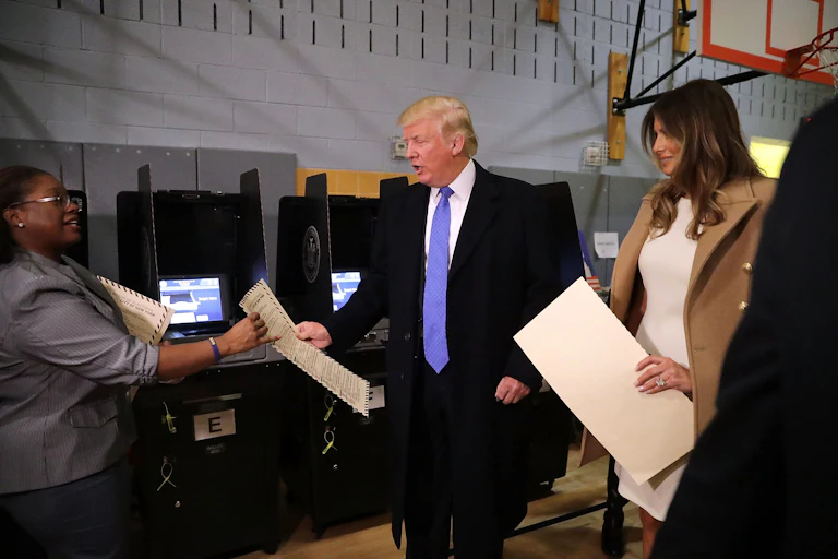 Donald Trump, center, and his wife Melania, right, hand ballots to a poll worker, left in a recreational facility with a basketball hoop visible in the background.