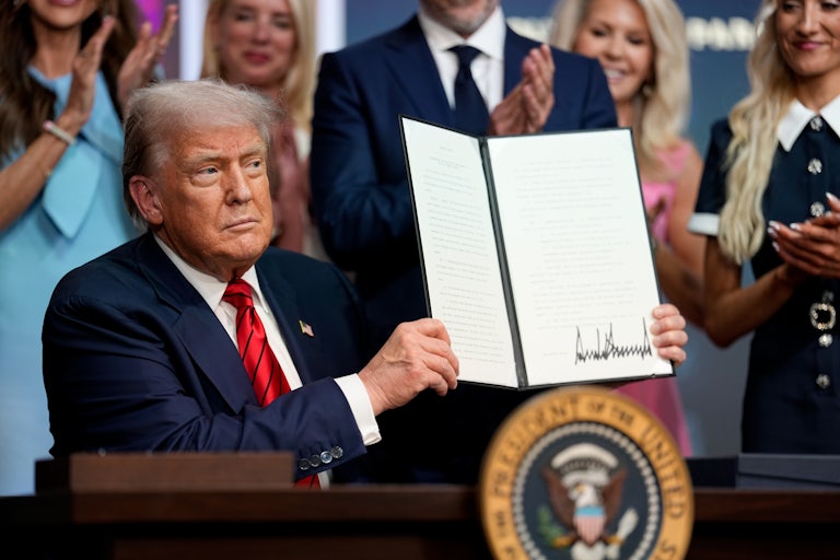 Donald Trump holds up a signed order seated at his desk in the White House while others stand behind him clapping.