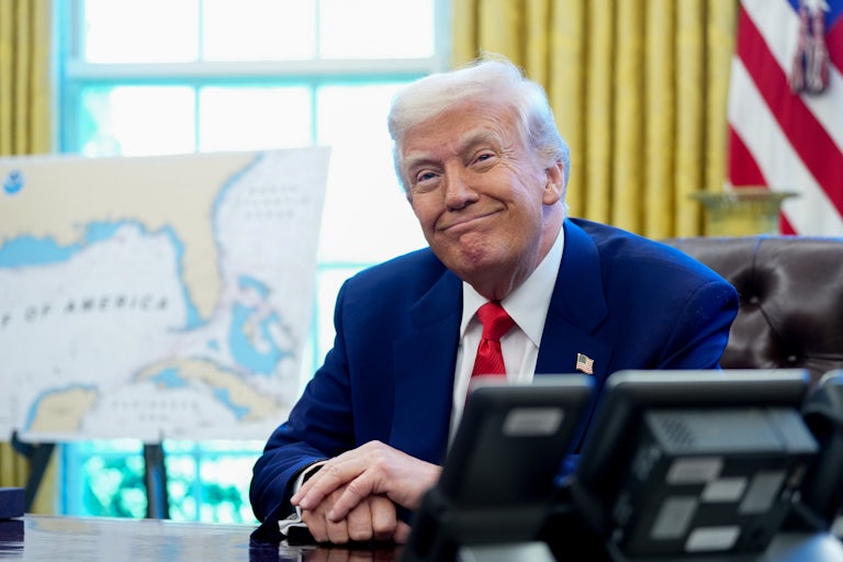Donald Trump smiles while seated at his desk in the White House. A map behind him, out of focus, shows the newly renamed "Gulf of America."