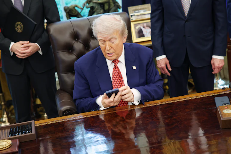 President Donald Trump looks at his phone while sitting at his desk in the Oval Office of the White House.