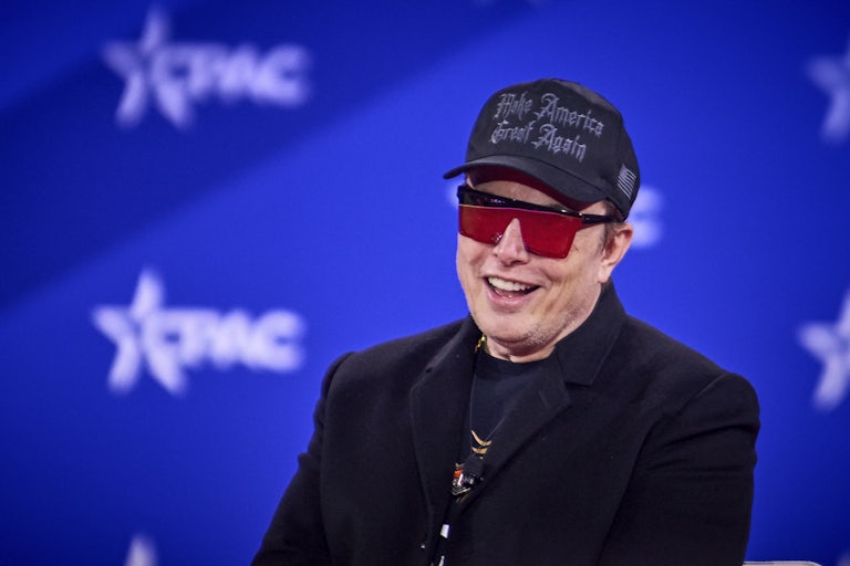 Elon Musk smiles at CPAC wearing absurd black and red sunglasses and a black MAGA cap.