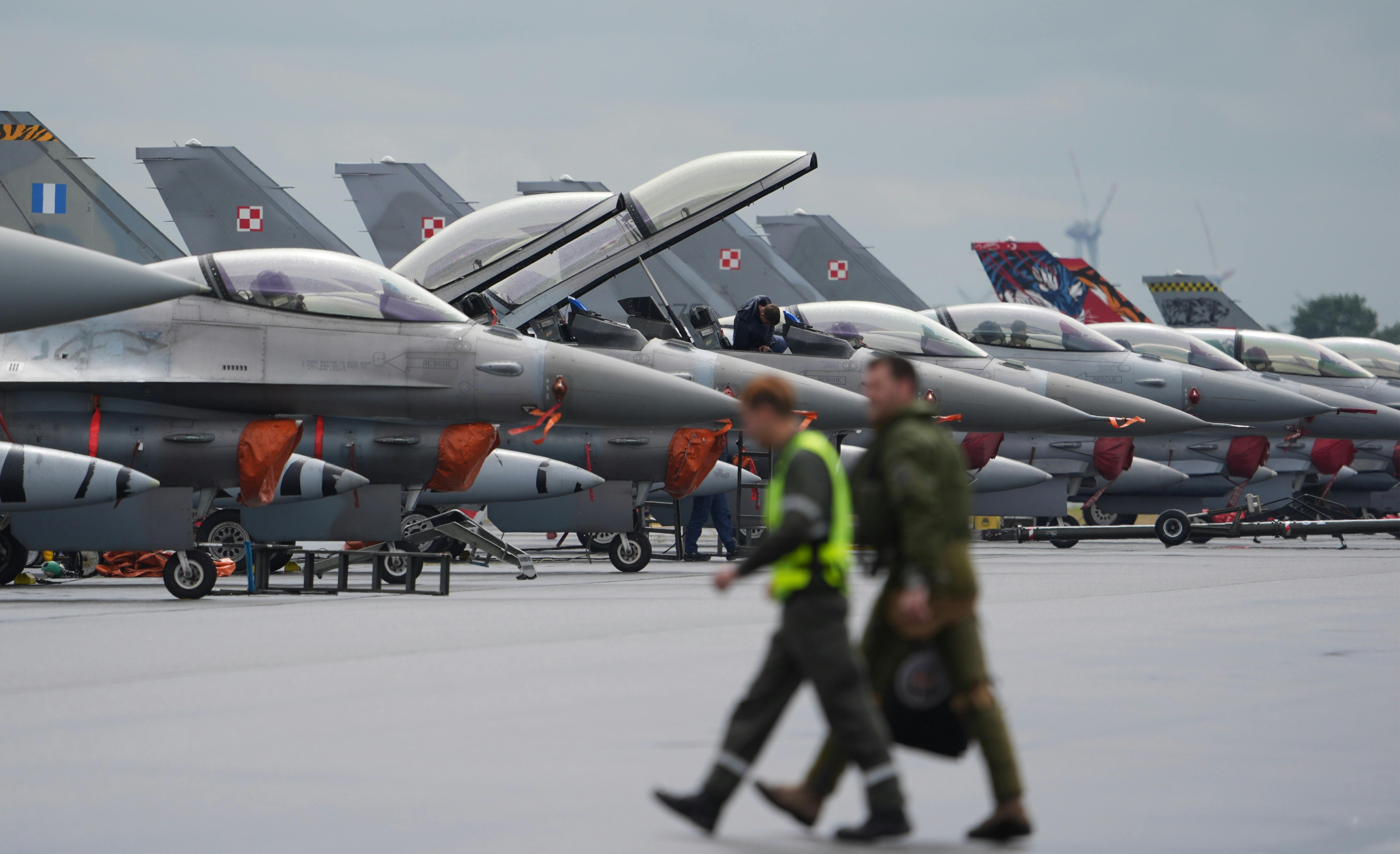 Two people in tactical uniforms walk in front of a line of fighter jets.