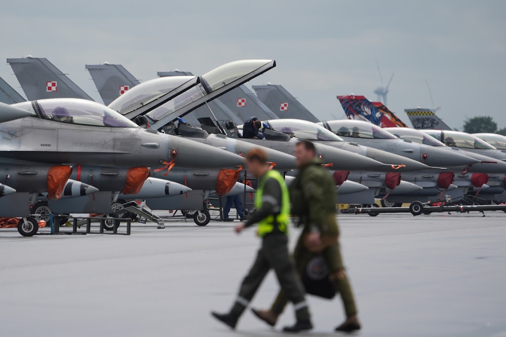 Two people in tactical uniforms walk in front of a line of fighter jets.