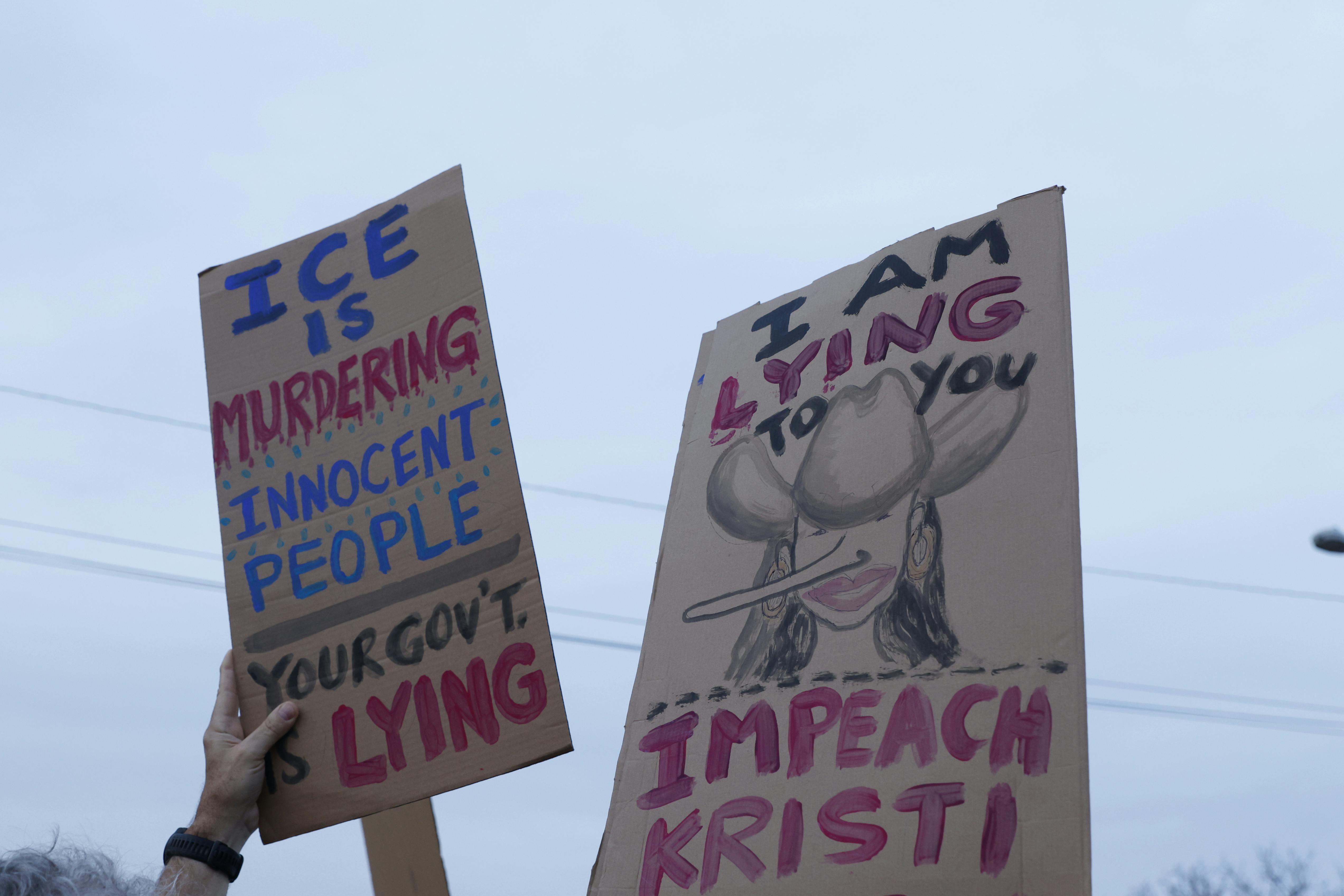 People hold up a sign that says, "ICE IS murdering innocent people. Your gov't is lying" and another that has a drawing of Kristi Noem with a cowboy hat and a Pinocchio nose that says, "I am lying to you. Impeach Kristi"