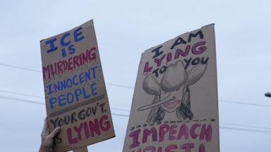 People hold up a sign that says, "ICE IS murdering innocent people. Your gov't is lying" and another that has a drawing of Kristi Noem with a cowboy hat and a Pinocchio nose that says, "I am lying to you. Impeach Kristi"