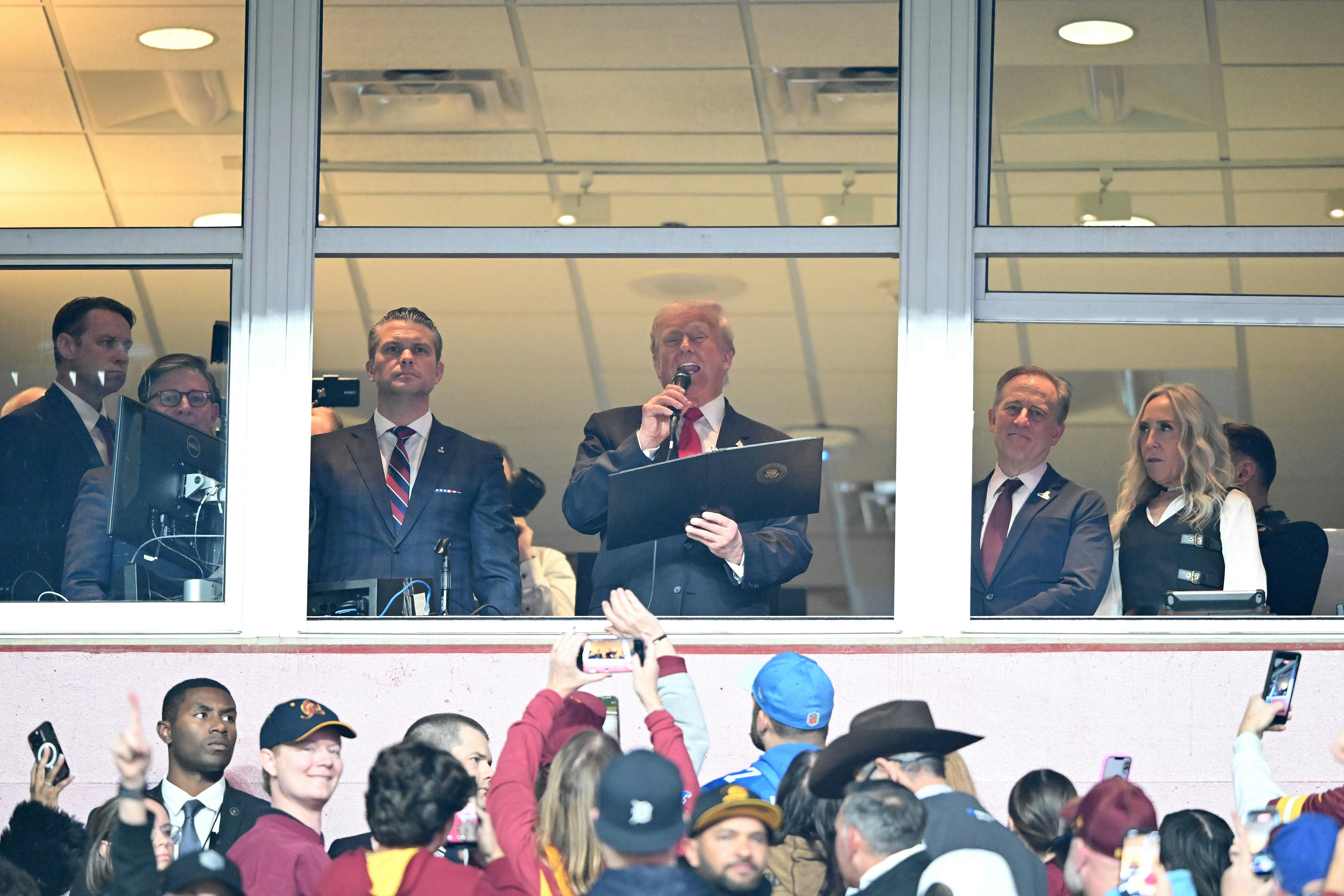 Donald Trump reads off a piece of paper into a microphone at the Commanders NFL game.