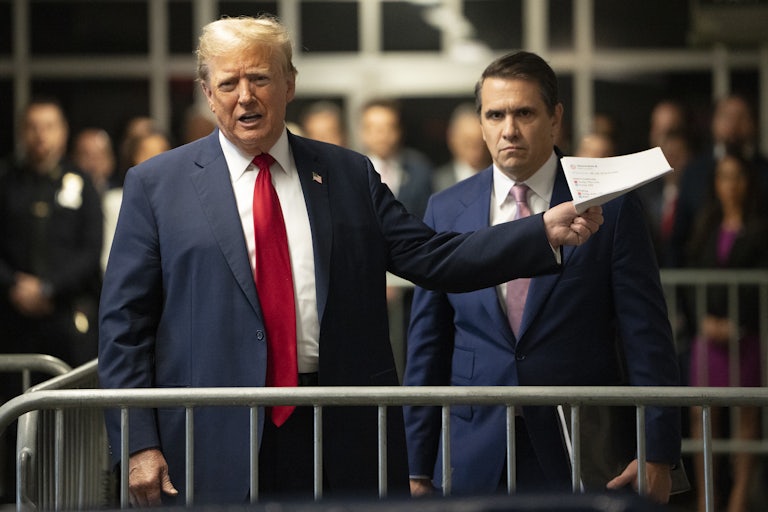 Donald Trump speaks and holds a stack of papers out in his left hand, standing in front of a metal barricade. Todd Blanche stands behind him. Others in the background are blurry.