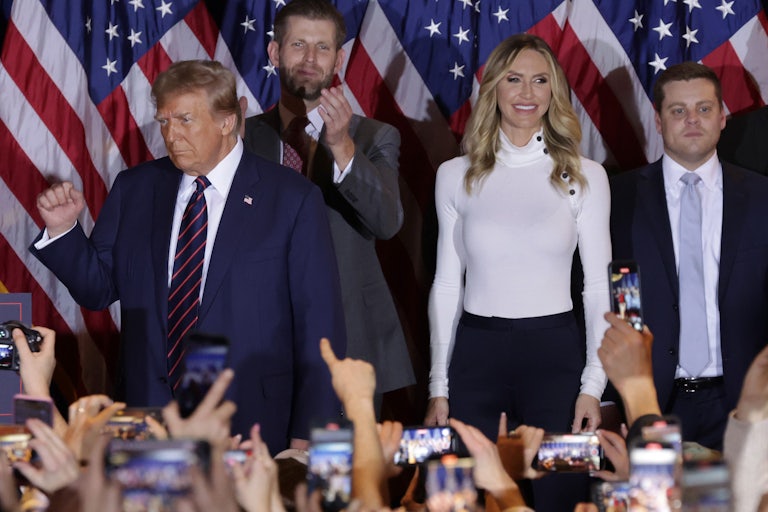 Donald Trump, Lara Trump, and Eric Trump stand on a stage wiith several U.S. flags behind them. You can see phones and hands raised in the crowd below them.