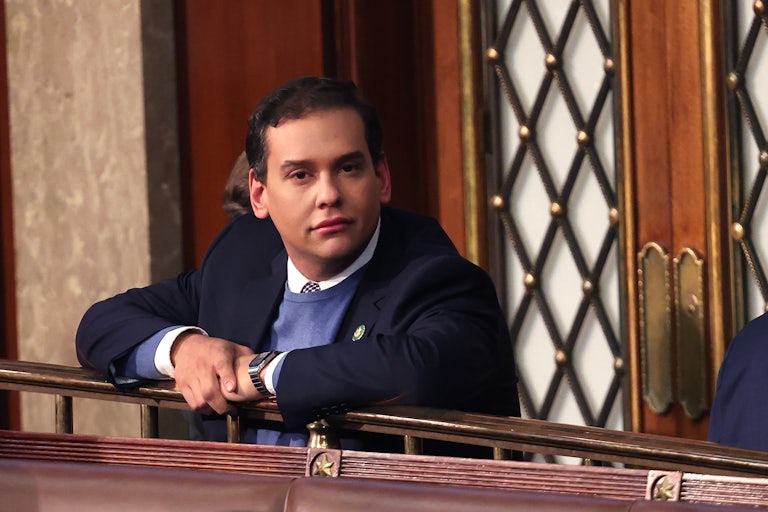 Representative George Santos leans over a railing in the House chamber and looks directly at the camera