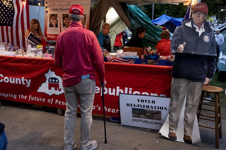 A man stands at a red Republican Party booth. A lifesize carboard cutout of Donald Trump gives a thumbs up.
