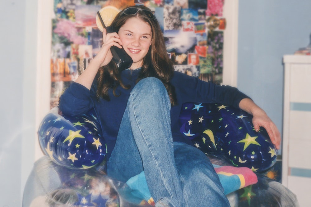 Teen wearing Y2K fashion, 2000s style, inside teenage bedroom, talking on cordless telephone, sitting on an inflatable chair, wearing flared denim jeans.