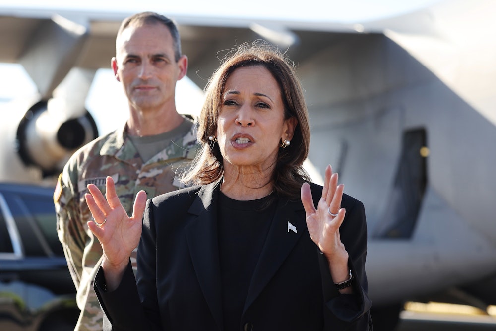 Kamala Harris gestures while speaking while a person in a camouflage uniform stands behind her.