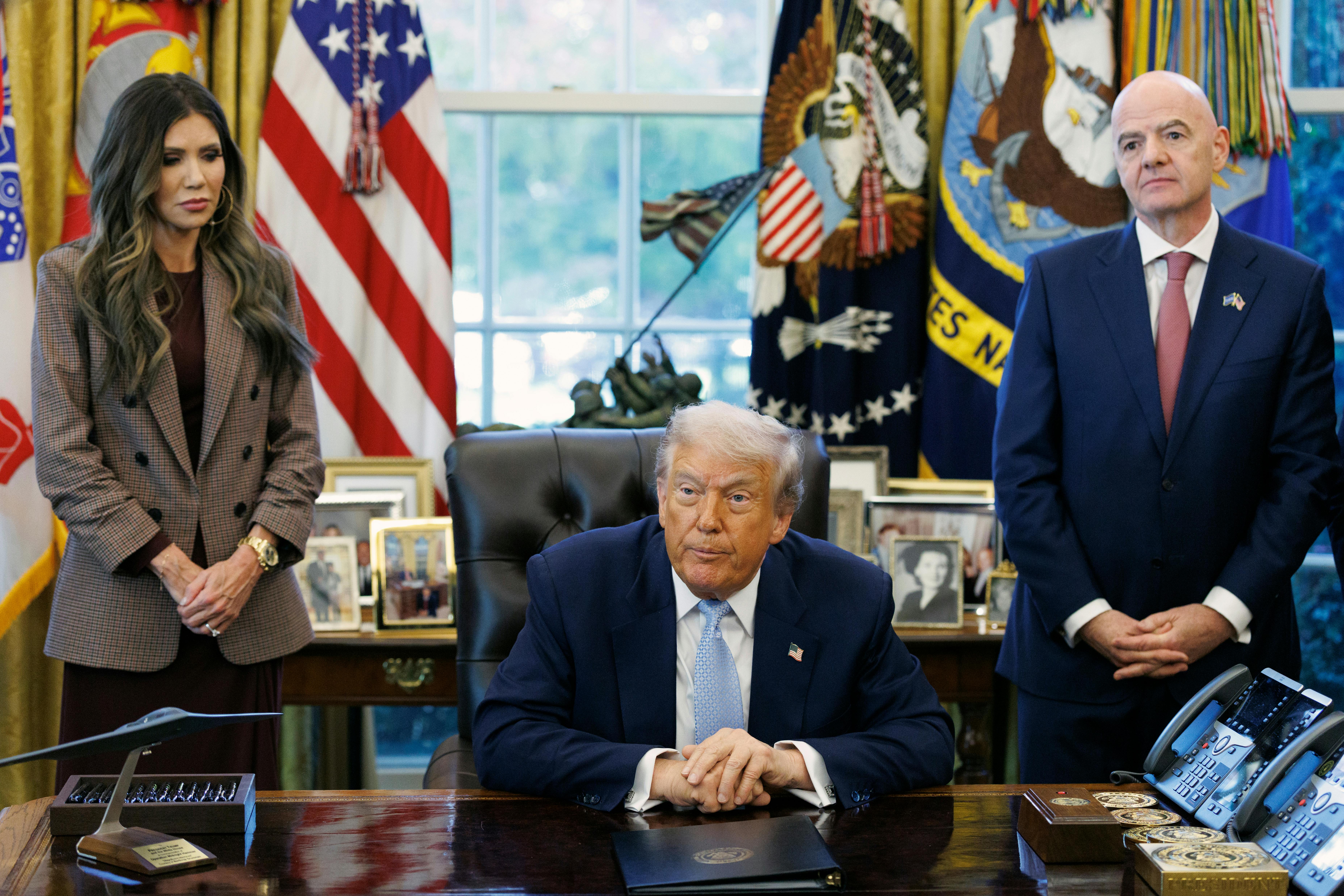 Homeland Security Secretary Kristi Noem and FIFA President Gianni Infantino stand on either side of Donald Trump, who is seated at his desk in the Oval Office of the White House.
