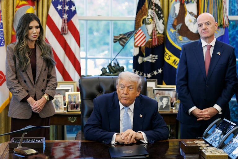 Homeland Security Secretary Kristi Noem and FIFA President Gianni Infantino stand on either side of Donald Trump, who is seated at his desk in the Oval Office of the White House.