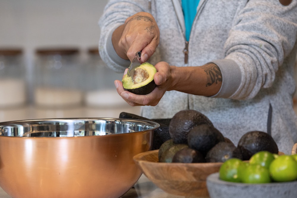 A person's arms are seen as the individual cuts into half of an avocado to remove the pit, admit a kitchen with bowls filled with avocados and limes.