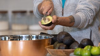 A person's arms are seen as the individual cuts into half of an avocado to remove the pit, admit a kitchen with bowls filled with avocados and limes.