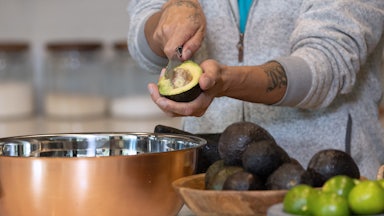 A person's arms are seen as the individual cuts into half of an avocado to remove the pit, admit a kitchen with bowls filled with avocados and limes.
