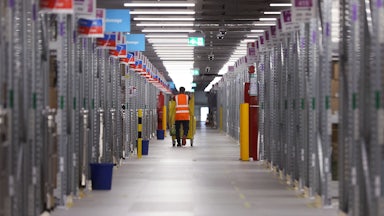 A worker in an orange safety vest pushes a cart of packages at an Amazon warehouse.