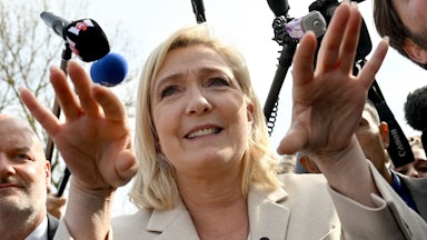 Marine Le Pen gesticulates while speaking, with microphones in her face.