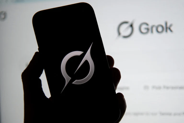 A hand holds a phone with the Grok logo while a screen in the background shows the Grok.com homepage.