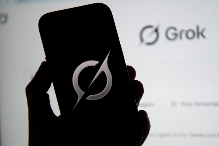A hand holds a phone with the Grok logo while a screen in the background shows the Grok.com homepage.