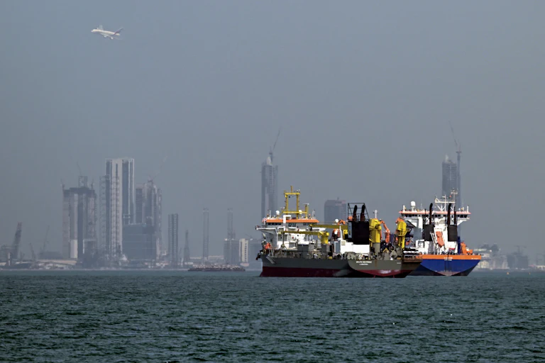 Two commercial ships sit in waters off the coast of Dubai, with towers in the background.