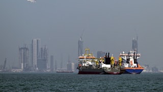 Two commercial ships sit in waters off the coast of Dubai, with towers in the background.