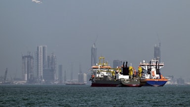 Two commercial ships sit in waters off the coast of Dubai, with towers in the background.