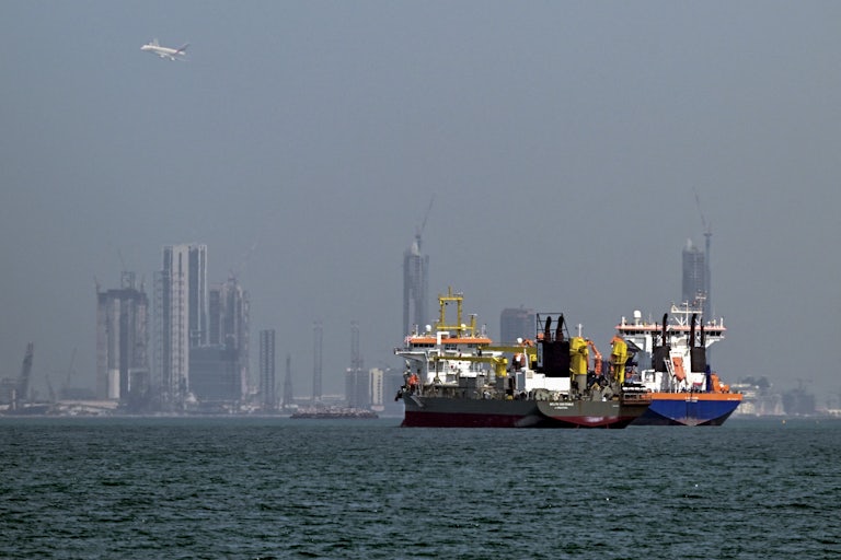 Two commercial ships sit in waters off the coast of Dubai, with towers in the background.