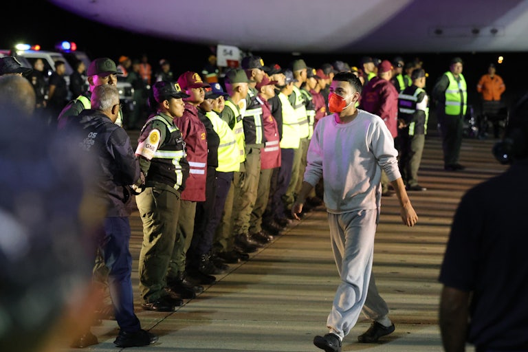 A man wears a gray sweatshirt, gray sweatpants, and a red face mask. He walks away from the plane as a line of airport workers and police officers watch him.