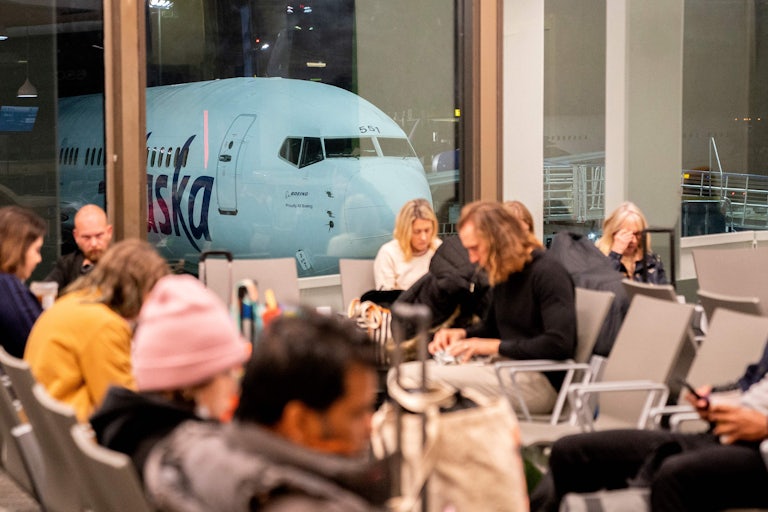 Travelers wait in the terminal as an Alaska Airlines plane can be seen from the window.