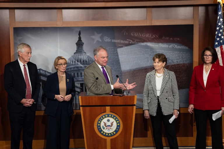 Senator Tim Kaine speaks at a podium about ending the shutdown. He is flanked by Senators Angus King, Maggie Hassan, Jeanne Shaheen, and Catherine Cortez Masto