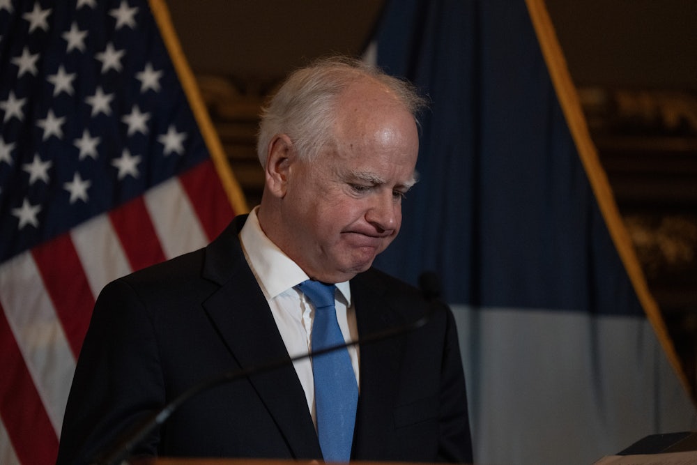 Tim Walz frowns, looking down, with an American flag in back of him.